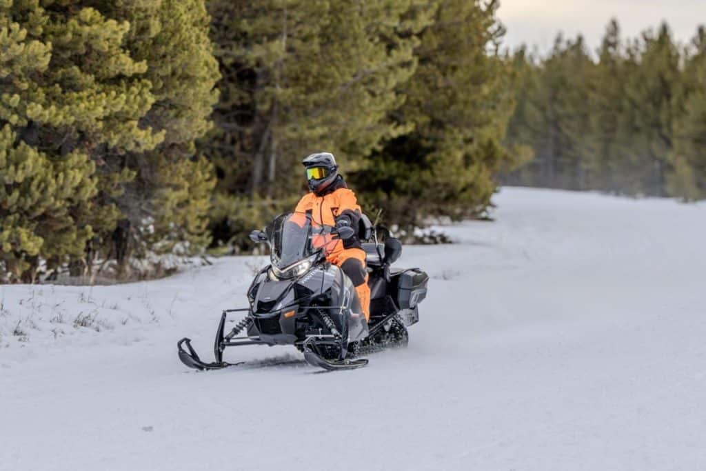 Rider wearing bright orange winter gear operating a black Arctic Cat Pantera snowmobile on a snowy forest trail surrounded by pine trees.