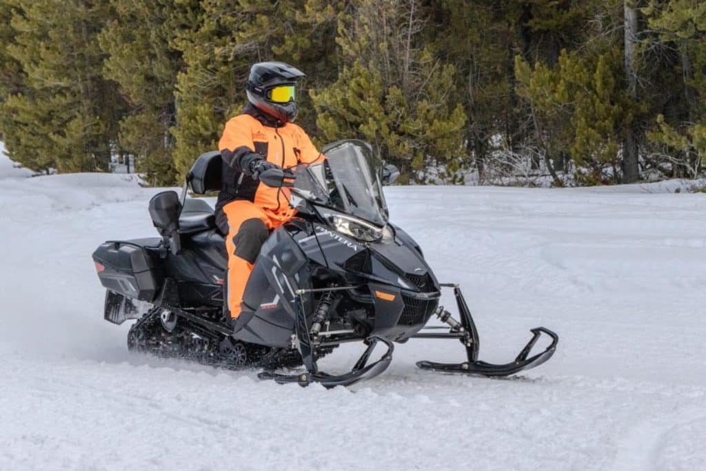 Rider in bright orange snow gear driving a black Arctic Cat Pantera touring snowmobile through snowy terrain surrounded by pine trees.