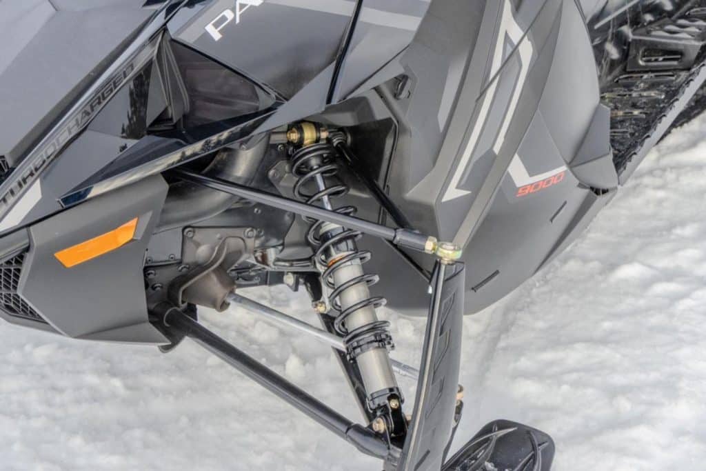 Close-up of the front suspension and ski assembly on a black Arctic Cat snowmobile, showing the shock absorber, steering arms, “Turbocharged” branding, and 9000 side panel details on snow.