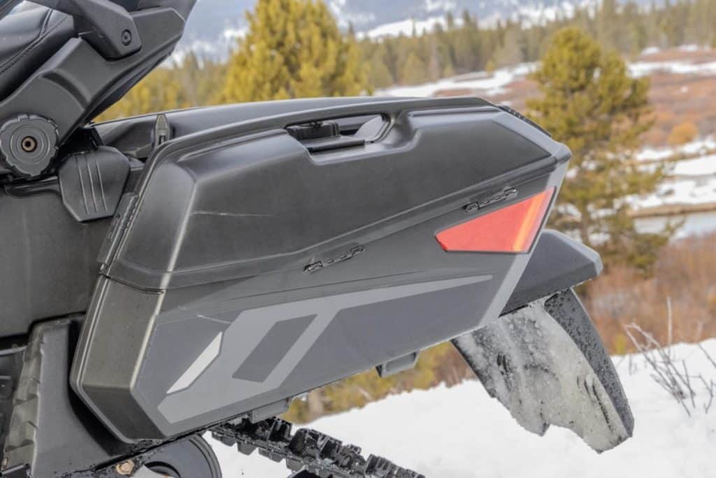 Close-up of a black snowmobile rear cargo storage box and taillight mounted above the track, with snowy terrain and trees in the background.