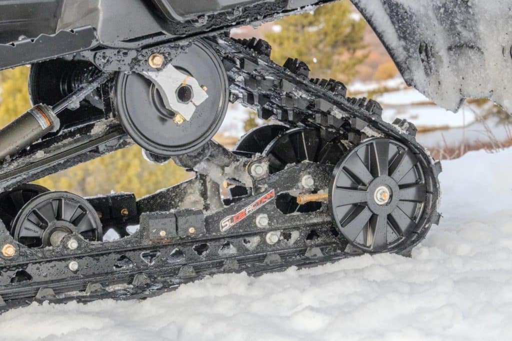 Close-up of a snowmobile rear suspension and track system with black idler wheels, shock absorber, and aggressive track lugs cutting through snow on a winter trail.