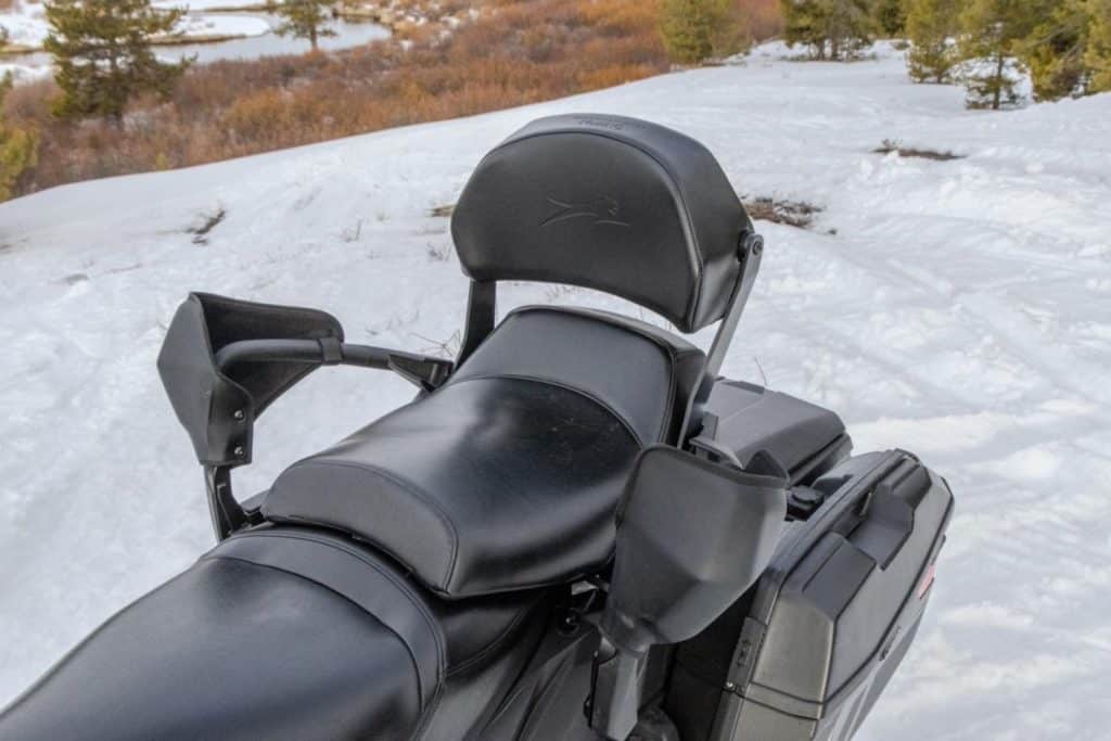 Passenger seating area of an Arctic Cat snowmobile featuring a padded backrest, hand guards, and rear cargo storage cases in a snowy outdoor setting.
