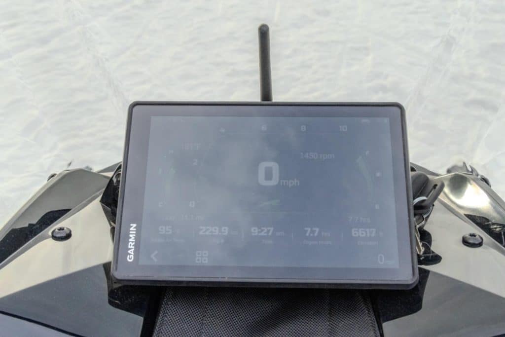 Close-up rider view of a Garmin touchscreen display mounted on an Arctic Cat snowmobile dashboard, showing speed, RPM, trip data, and engine information on snowy terrain.