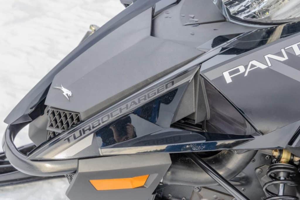 Close-up of a black Arctic Cat snowmobile front side panel with “Turbocharged” branding, angular bodywork, front suspension, and headlight visible on a snowy background.
