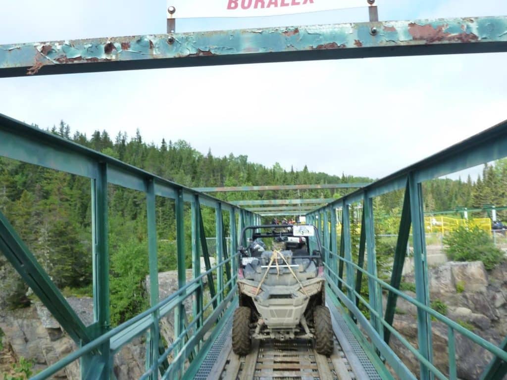 Side-by-side ATV crossing a narrow metal bridge above a rocky canyon, surrounded by a dense green forest under a clear sky.
